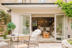 A cozy outdoor seating area with wicker furniture, a wooden table, and a glass door leading to an interior space with a kitchen and dining area.