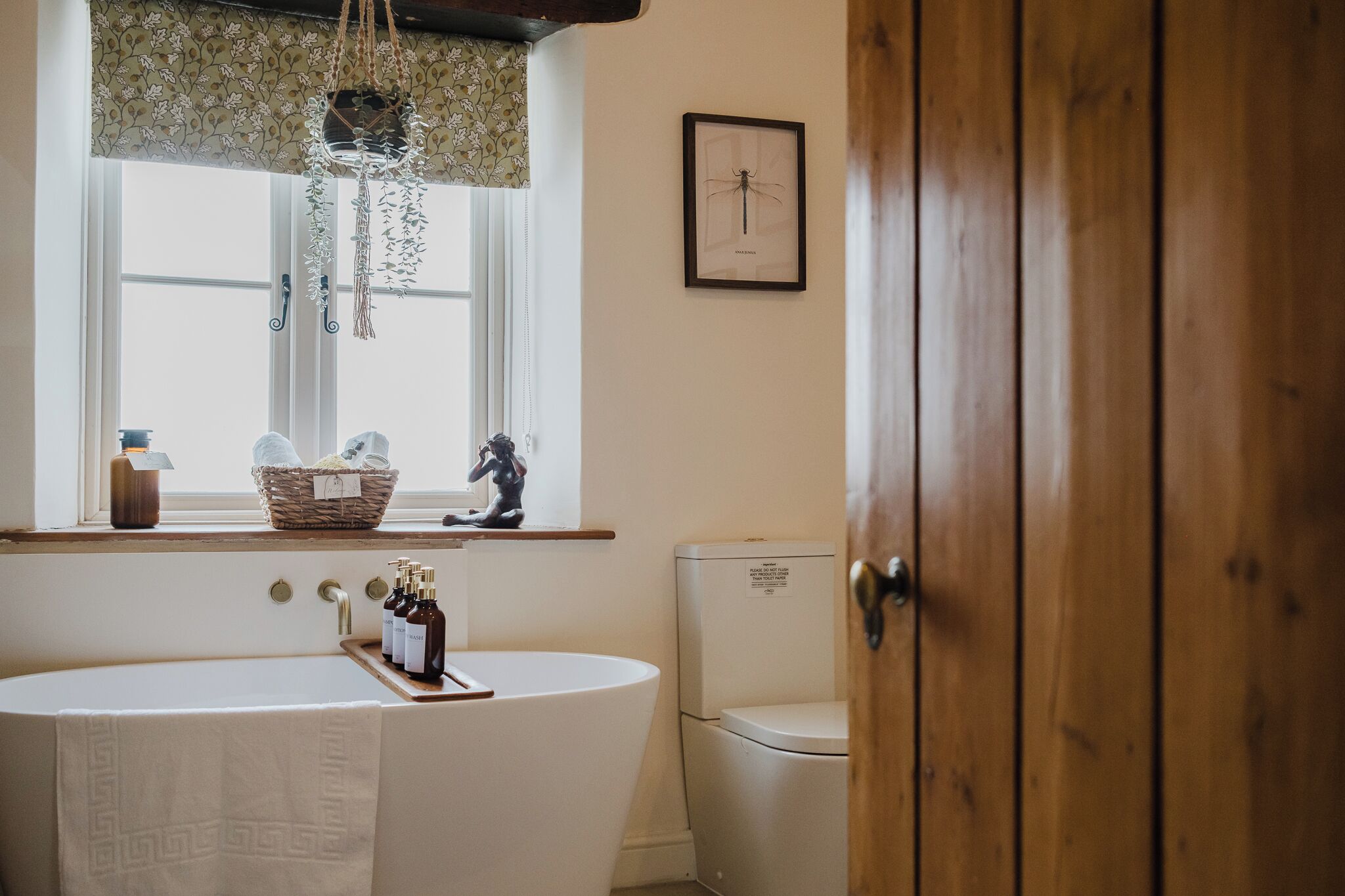 Ein Badezimmer mit freistehender Badewanne, Toilette, einem Fenster mit Wandbehang aus Makramee und einem Insektenbild im Bilderrahmen an der Wand.