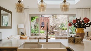 A modern kitchen with a central island, pendant lights, and a view of a stone wall and garden. - Beautiful Cottage in Cirencester Centre (Cirencester)