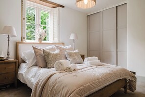 A neatly made bed with a beige comforter, two bedside lamps, a window with a view of greenery, and a large wardrobe.