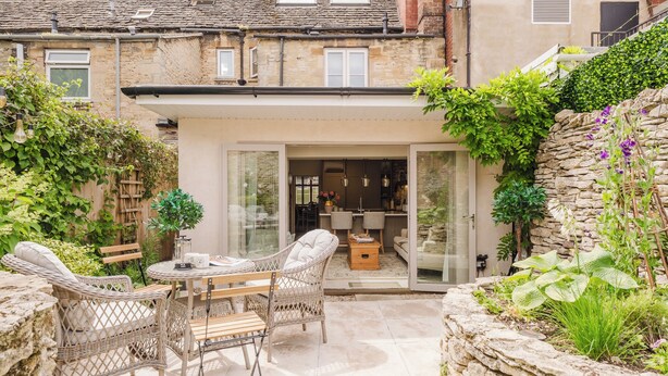A patio with wicker furniture, a stone wall, and a glass door leading to a room with a dining area. - Beautiful Cottage in Cirencester Centre (Cirencester)