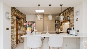 A kitchen with a stone wall, two bar stools, a white countertop, hanging pendant lights, and a clock on the wall. - Beautiful Cottage in Cirencester Centre (Cirencester)