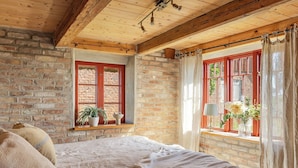 A rustic bedroom with exposed wooden beams, a brick wall, a bed with a white bedspread, and a window with red shutters.