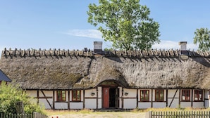 A traditional thatched-roof house with a wooden fence in front, a gravel path leading to the entrance, and a tree to the right.