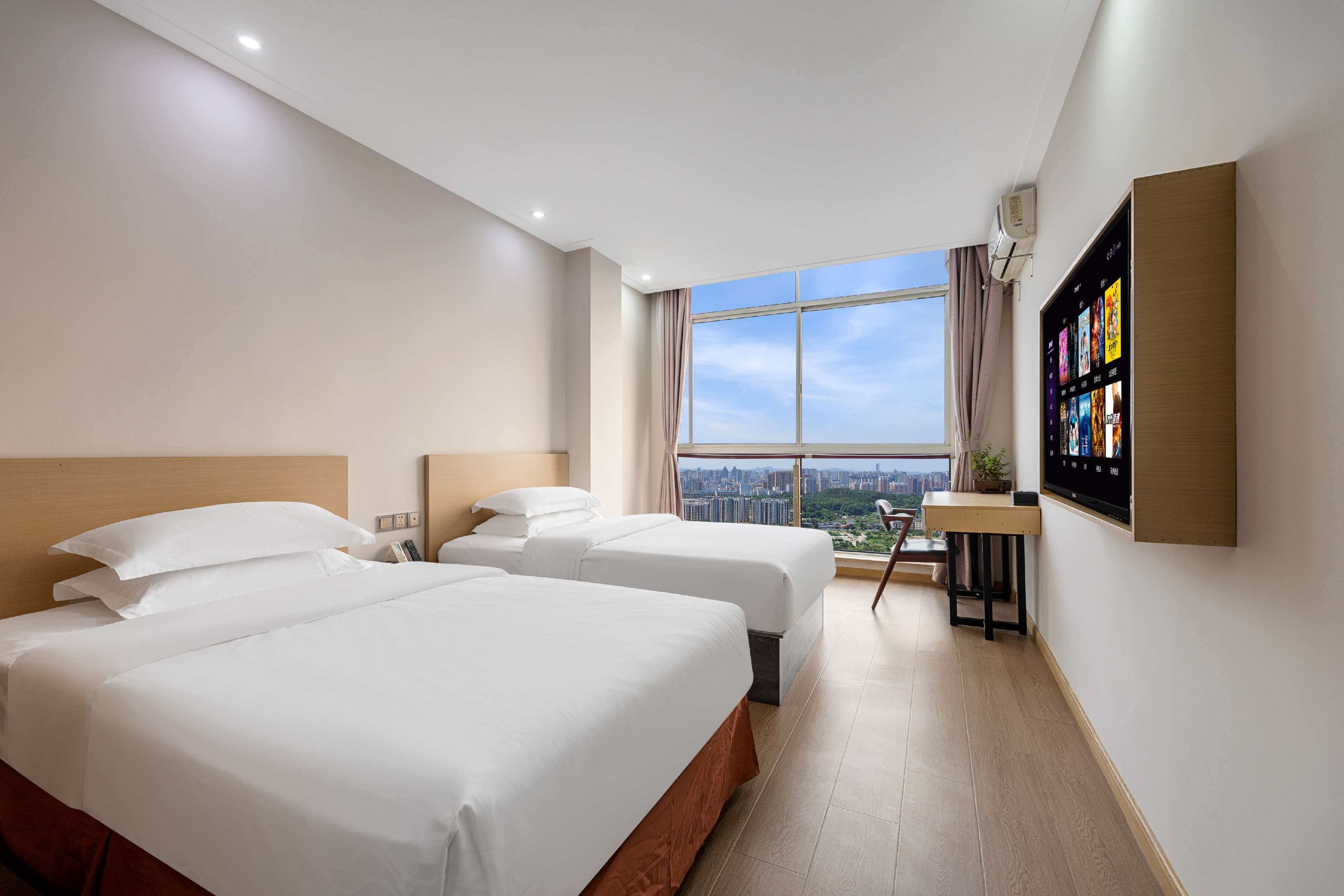 A hotel room with two beds, a desk, a TV, and a large window with a city view.