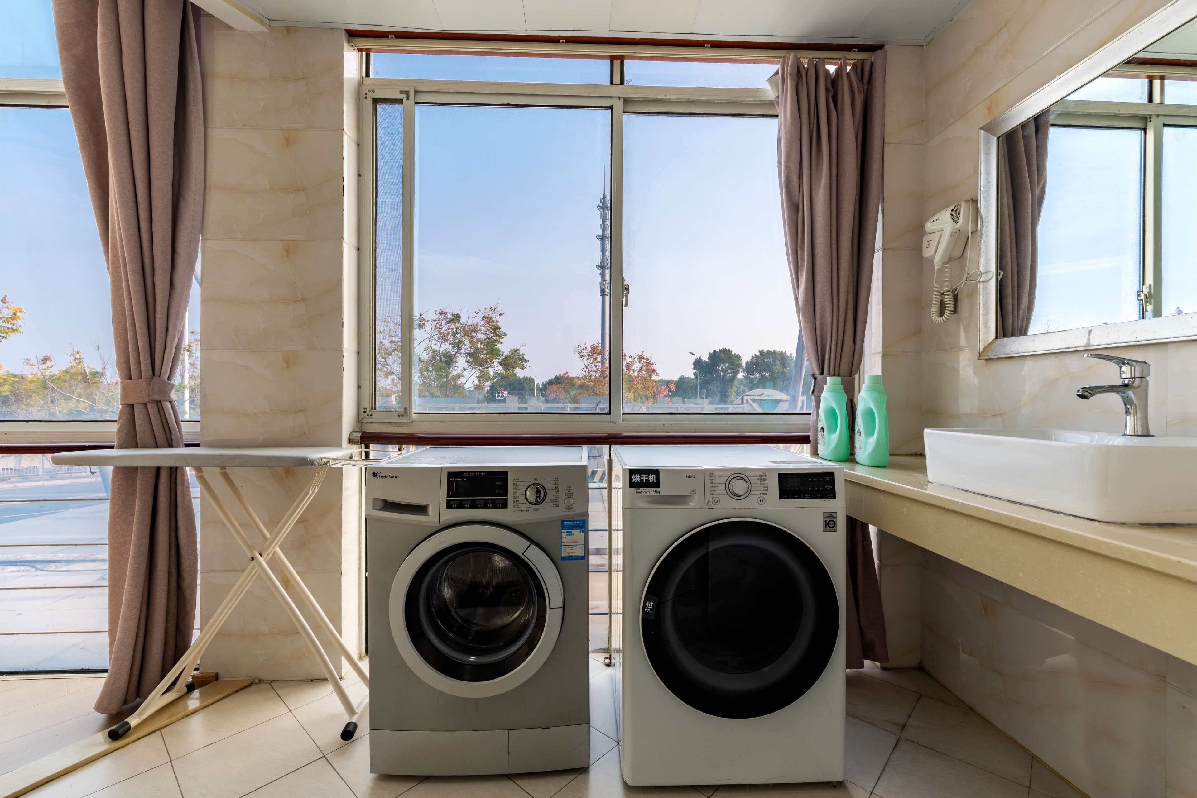 A compact hotel room with a washing machine, a dryer, a sink, and a window with curtains.