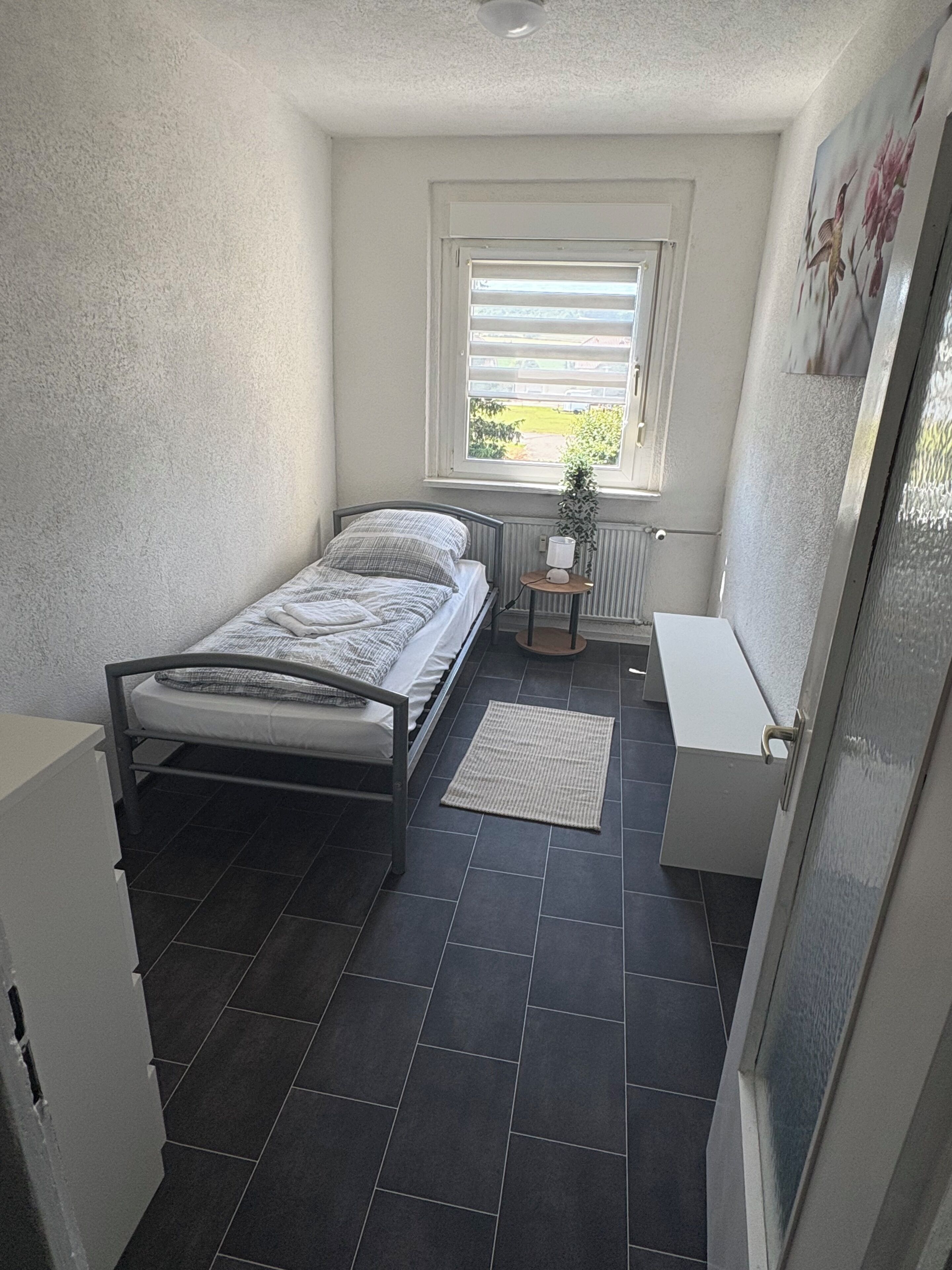 Une petite chambre à coucher, de taille réduite, avec un lit simple, un radiateur, une petite table avec une lampe et une fenêtre équipée de stores.