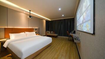 A hotel room with a large bed, a desk, a chair, a projector screen, and a wall-mounted TV.