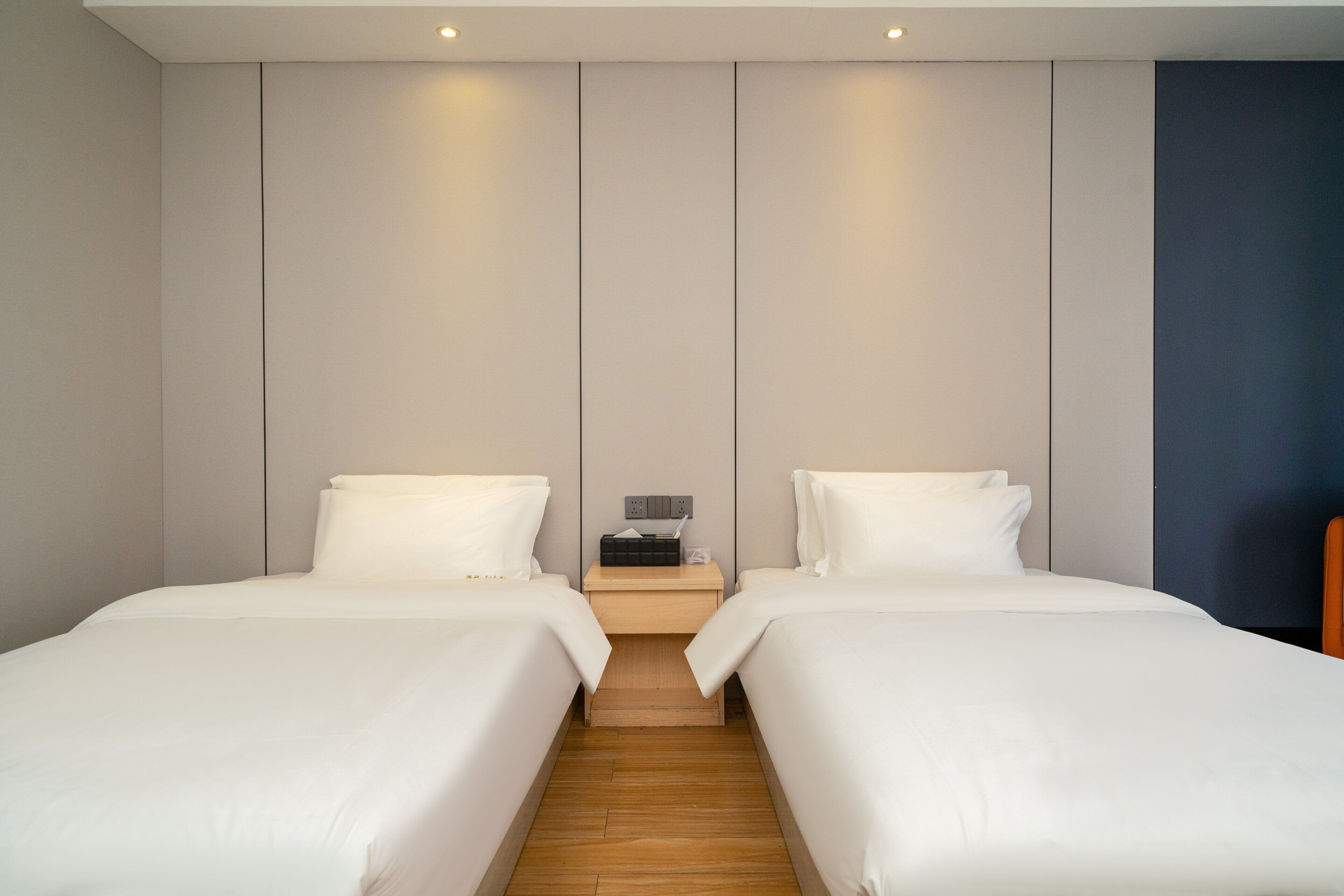 Habitación de hotel con dos camas, piso de madera, mesita de noche con un teléfono y una lámpara, y una pared con diseño de paneles.