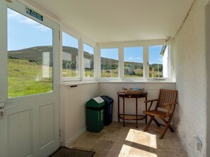 A room with a door marked 'Fire exit' and 'Keep clear,' a window with a view of a green landscape, a trash bin, a small table with a basket, and a wooden chair. - Strathnoon by Interhome (Tomatin)
