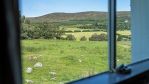 A view through a window of a grassy field with scattered rocks, a hill in the distance, and a house. - Strathnoon by Interhome (Tomatin)