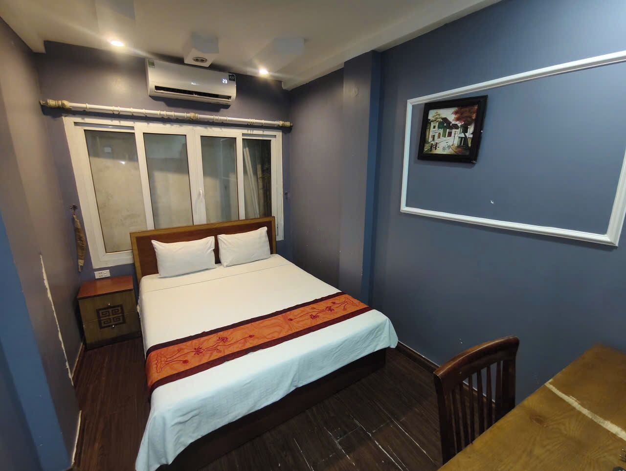 A bedroom with a bed, a wooden nightstand, a table, a chair, a window with curtains, and a wall-mounted picture.
