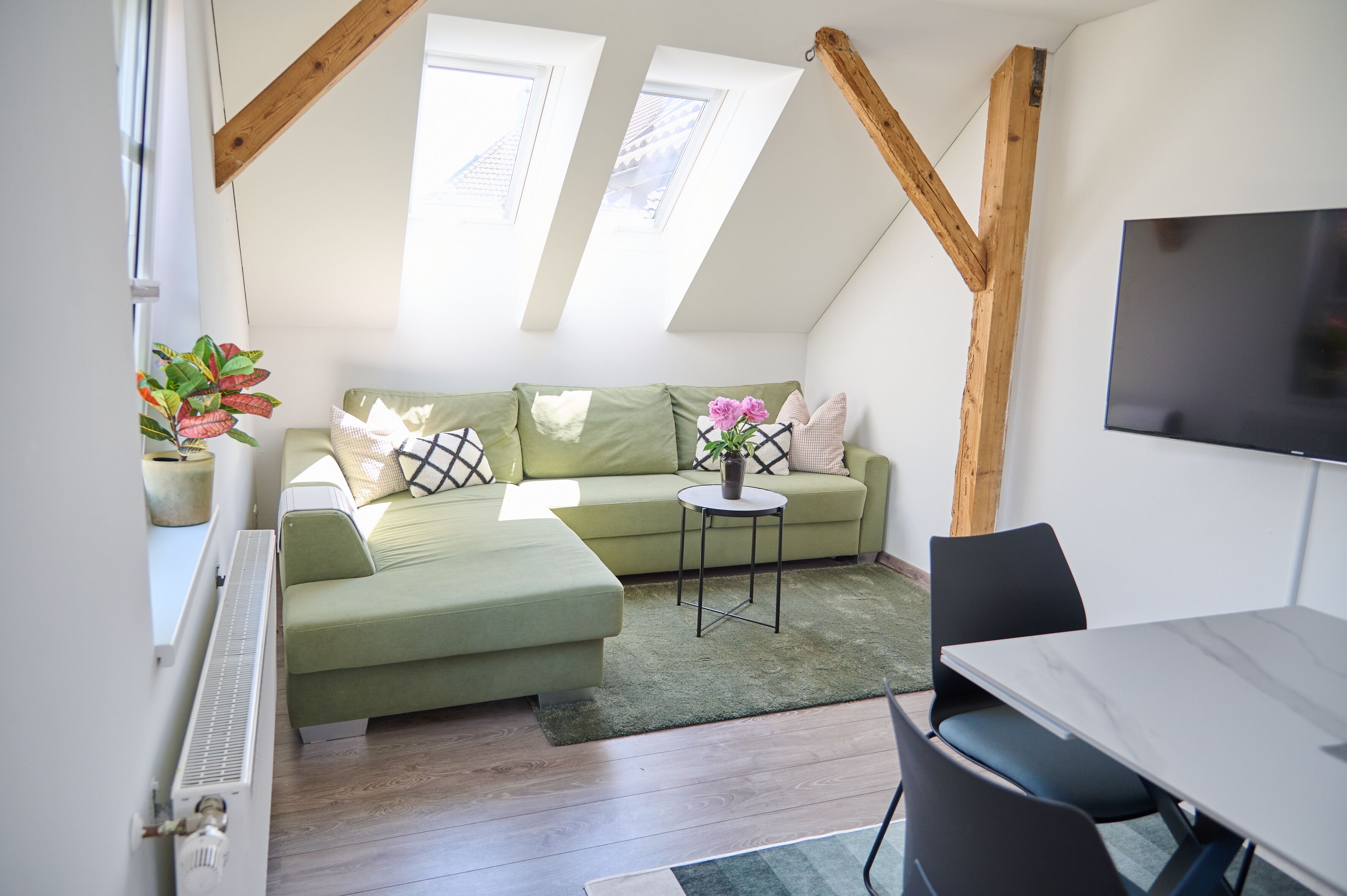 A cozy loft bedroom with a green sofa, a small round table, and a TV mounted on the wall.
