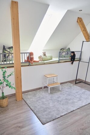 Una habitació amb sostre inclinat, una petita banca blanca, un fosc i petit moqueta, un suport de fusta per a plantes i una escala metàl·lica.