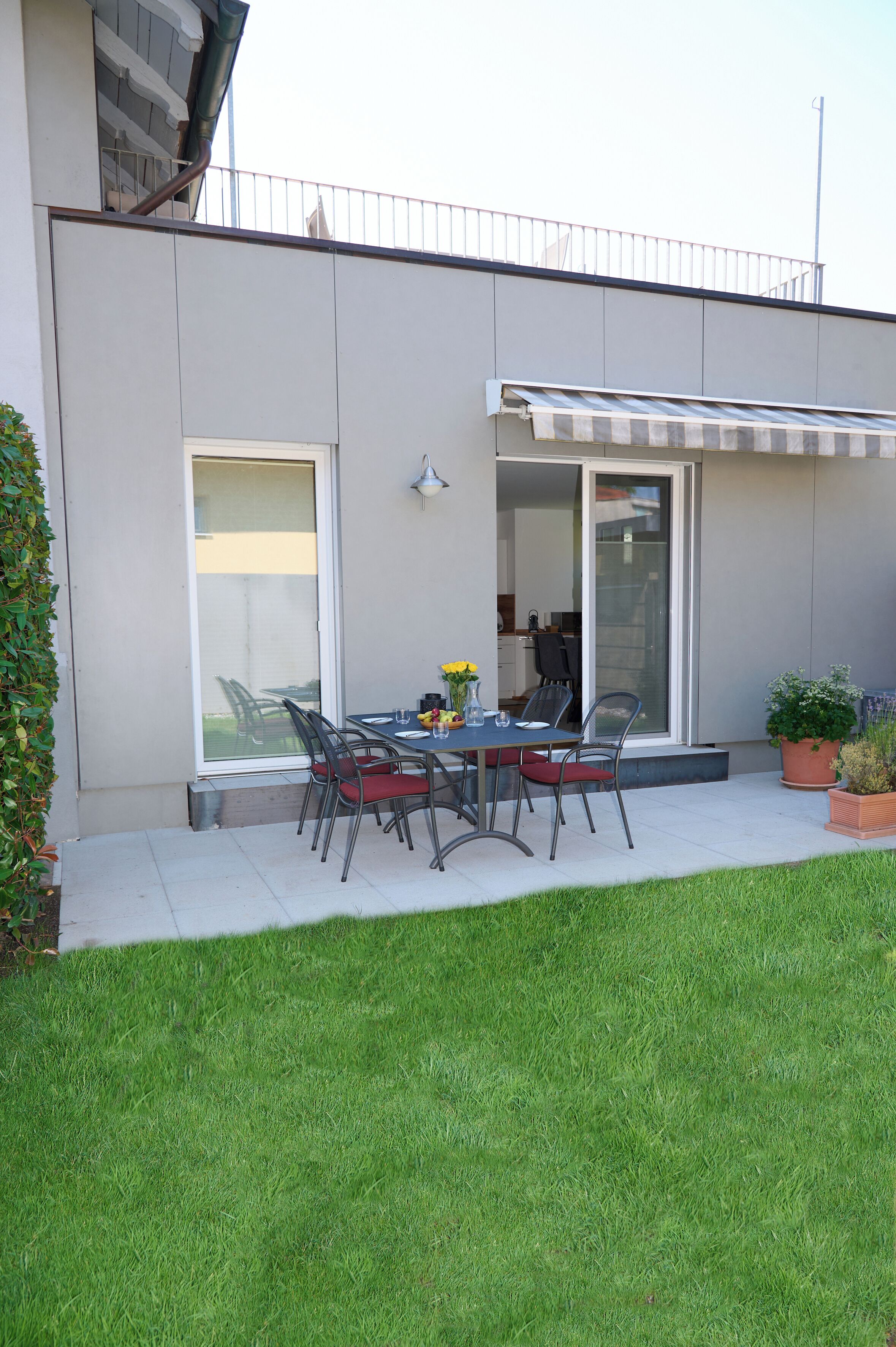 A modern outdoor dining area with a table and chairs, a glass door leading to a covered patio, and a well-maintained lawn.