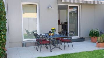 A modern outdoor dining area with a table and chairs, a glass door leading to a covered patio, and a well-maintained lawn.