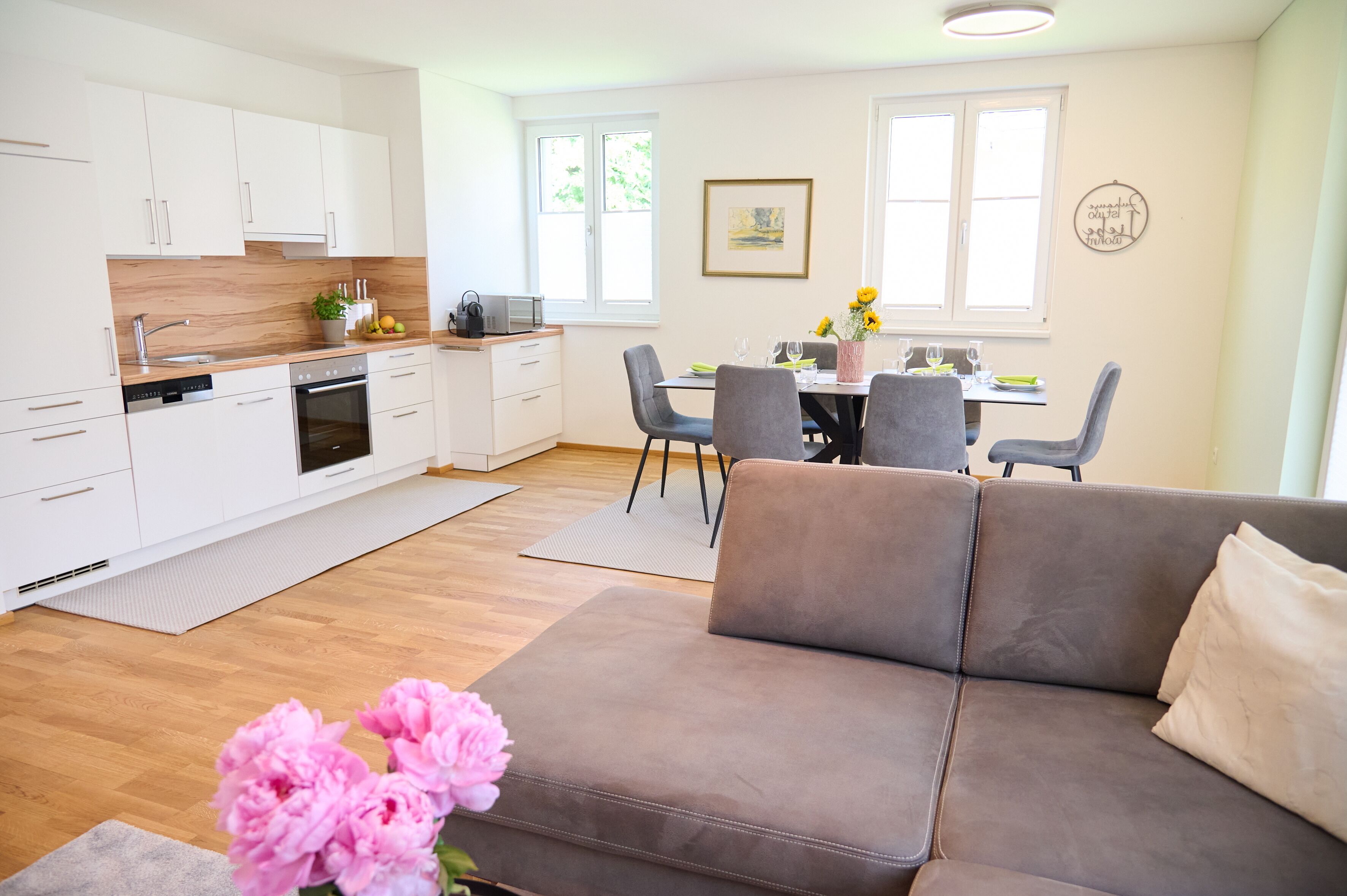 A modern kitchen with white cabinets, a dining area with a table set for four, and a grey sofa in the living area.