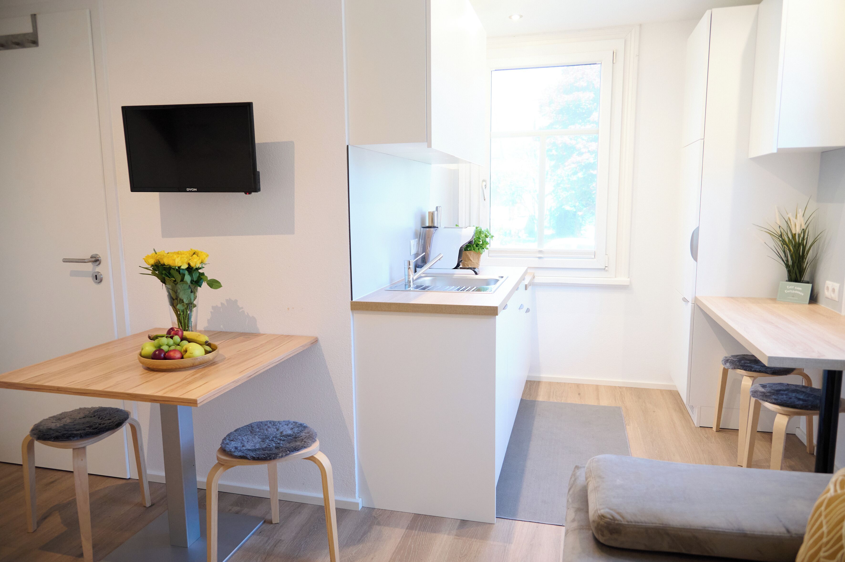 A compact kitchen with a dining area, a flat-screen TV mounted on the wall, and a window providing natural light.