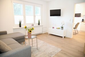 A modern living room with a grey sofa, a small round table with a vase of flowers, a white entertainment unit with a flat-screen TV, and a wooden floor. - Villa Rebgarten (Höchst)