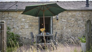 Un bâtiment en pierre avec une table et des chaises sur une terrasse, protégées par un parasol vert.