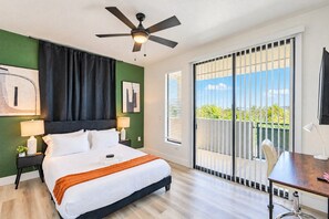 Habitación de hotel con cama, escritorio, silla, ventilador de techo y vista al exterior a través de una puerta corrediza.