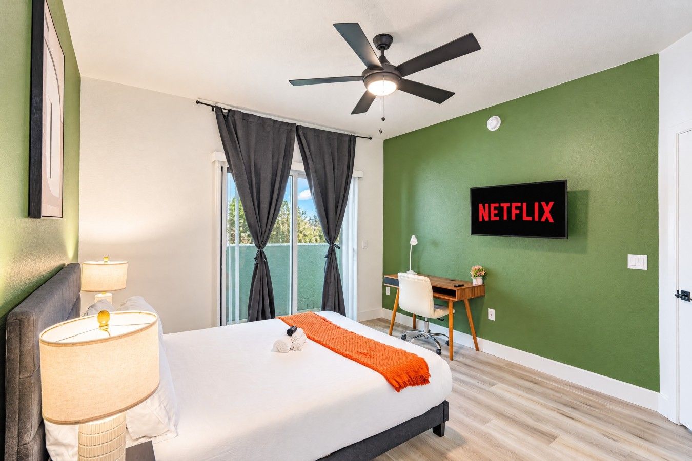 A hotel room with a bed, a desk, a ceiling fan, a TV displaying 'NETFLIX', and a window with curtains.