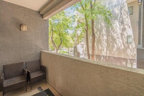 A balcony with a wicker bench, a textured wall, and a view of trees and buildings. - Glendale Stadium Retreat  Pool, Gym & Westgate AZ (Glendale)