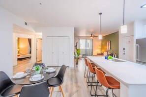 A modern kitchen with a central island, a dining area with a table set for four, and a separate living area with a sofa and a TV. - Glendale Stadium Retreat  Pool, Gym & Westgate AZ (Glendale)