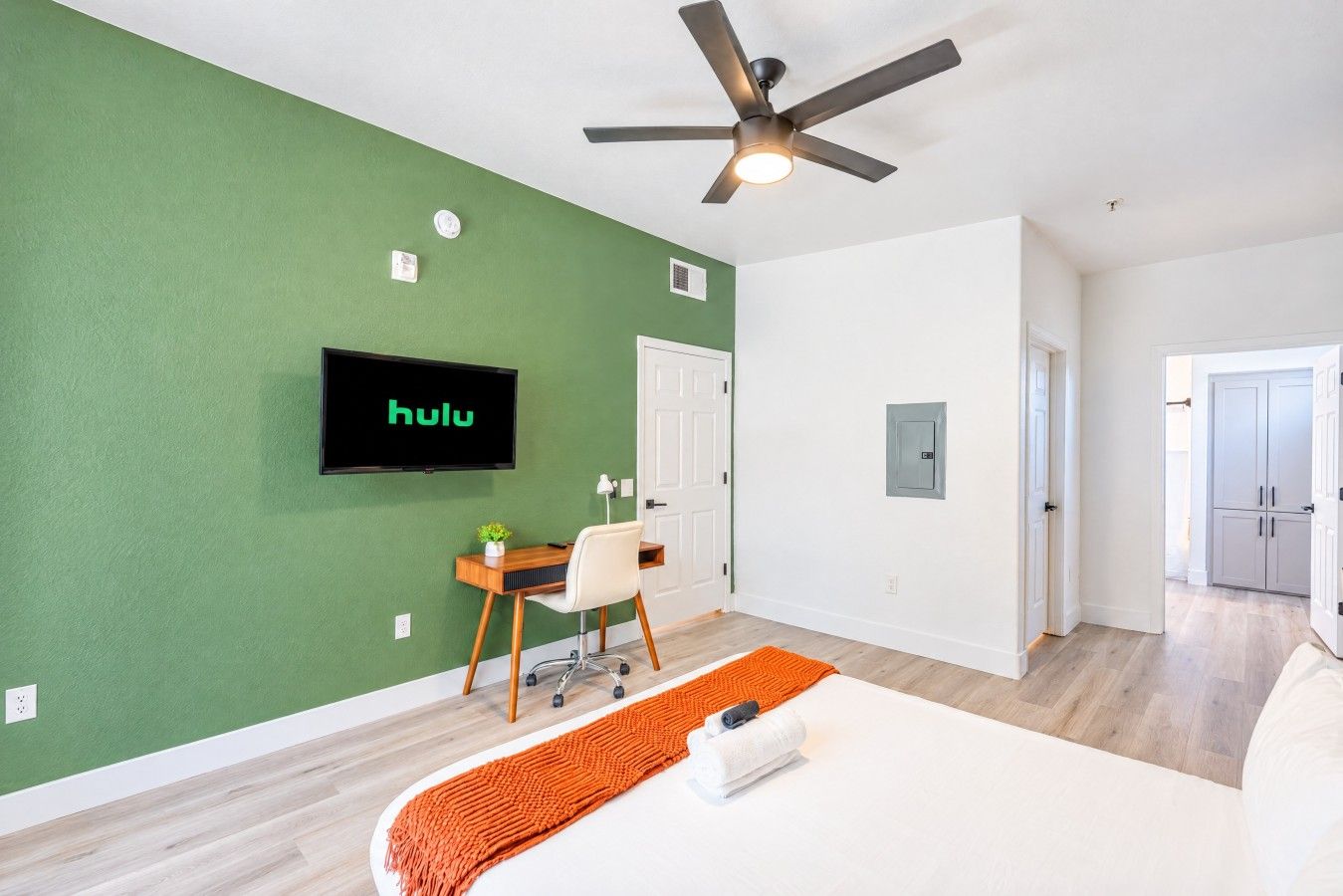 Ein modernes Zimmer mit grüner Wand, einem Deckenventilator, einem an der Wand befestigten Fernseher, einem Schreibtisch mit Stuhl und einem Bett mit orangefarbener Tagesdecke.