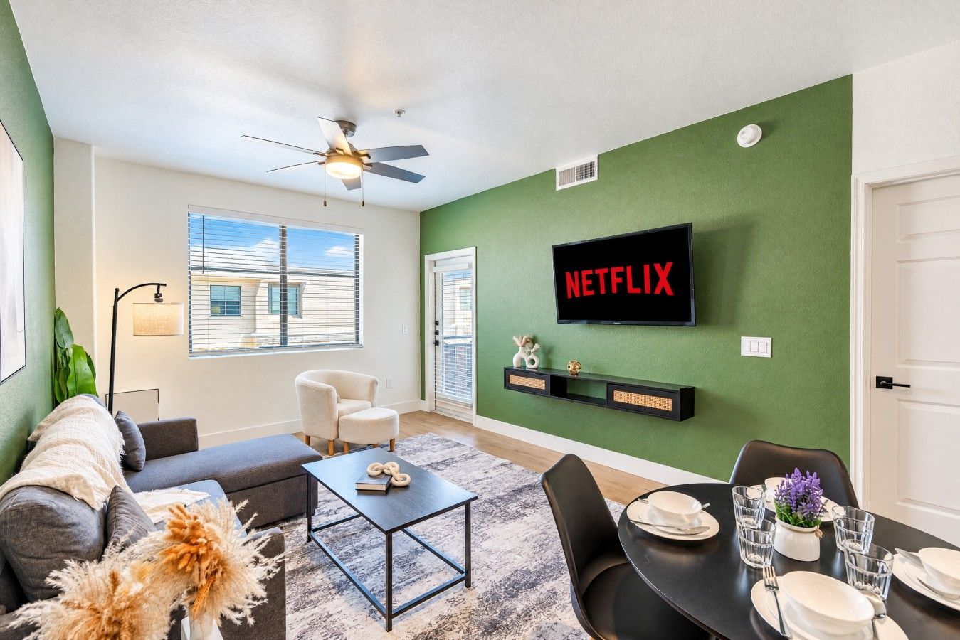 A living room with a green wall, a flat-screen TV mounted above a shelf, a ceiling fan, a sofa, a coffee table, and a dining area with a table set for two.