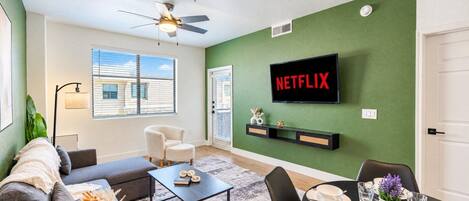 A living room with a green wall, a flat-screen TV mounted above a shelf, a ceiling fan, a sofa, a coffee table, and a dining area with a table set for two.