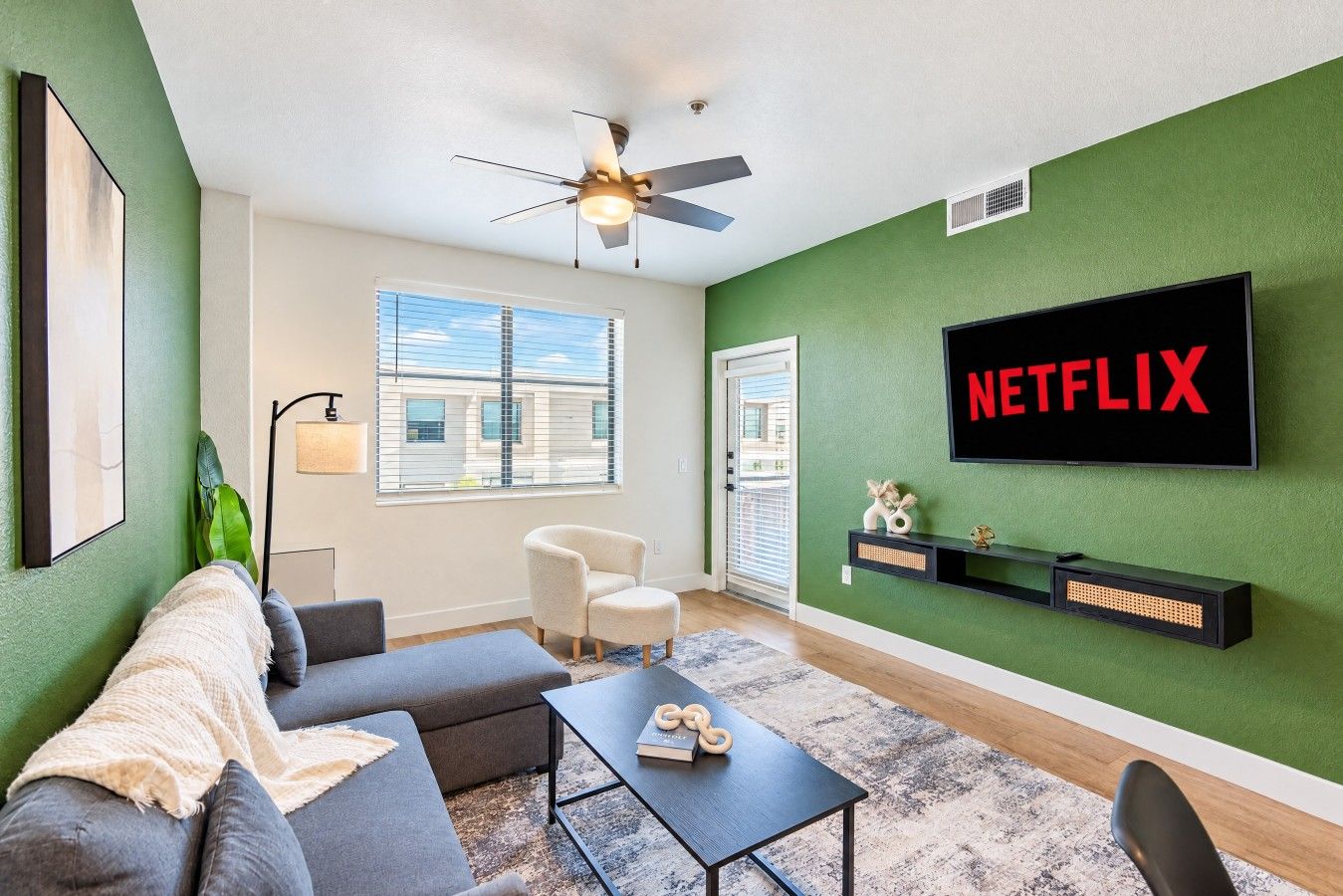 Un salon avec un mur vert, un ventilateur de plafond, un téléviseur à écran plat fixé au-dessus d’une étagère noire et une grande fenêtre avec des stores.