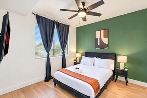 Ein Hotelzimmer mit Bett, Deckenventilator, Fernseher, Fenster mit Vorhängen, Nachttisch mit Lampe und grüner Wand.
