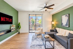 A living room with a flat-screen TV, a ceiling fan, a sofa, a coffee table, and a view through a glass door to the outside. - Stadium District Retreat Pool, Gym & Westgate (Glendale)