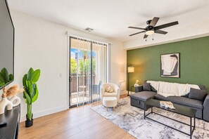 A living room with a sofa, a coffee table, a chair, a ceiling fan, and a sliding glass door leading to an outdoor area. - Stadium District Retreat Pool, Gym & Westgate (Glendale)
