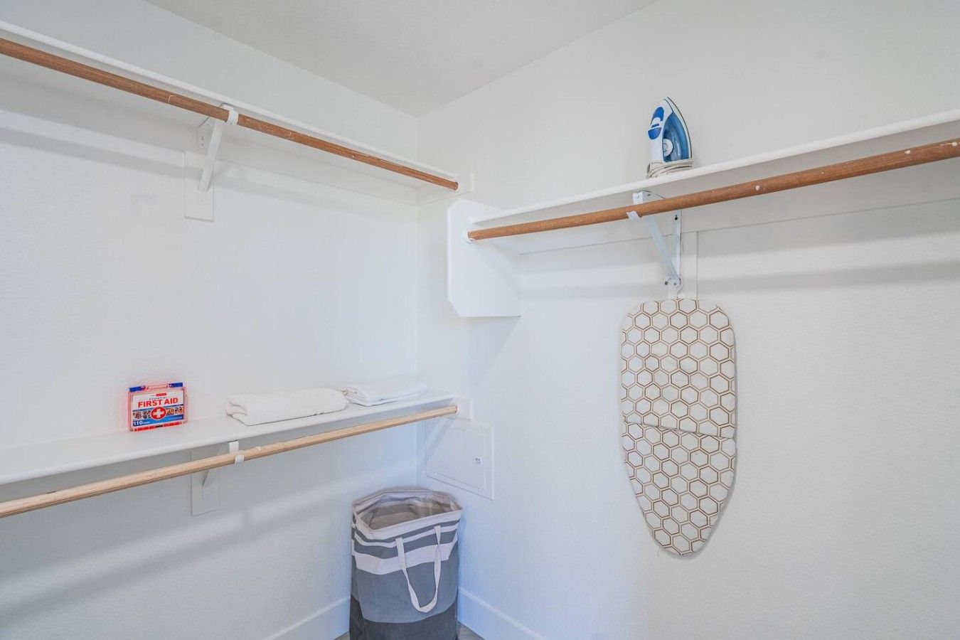 A laundry area with shelves, an ironing board, a first aid kit, and a trash bin.