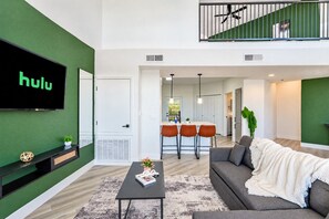 A modern living room with a large flat-screen TV, a sofa, a coffee table, and a kitchen area in the background. - Glendale Westgate Stay  Pool Gym WD Near Stadium (Glendale)