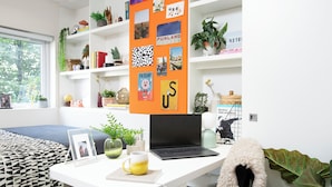 Quarto moderno com escrivaninha branca, laptop, fotografia emoldurada, caneca e estante com diversos objetos.
