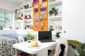 A modern room with a white desk, a laptop, a framed photo, a mug, and a bookshelf with various items. - Student Only Adam Street Gardens (Cardiff)