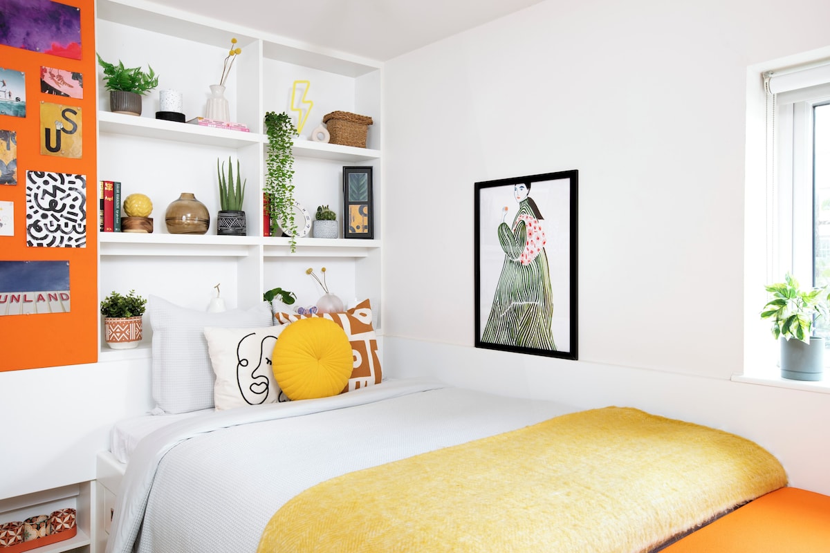 A bedroom with a bed, a window, a shelf with plants, and a framed picture on the wall.