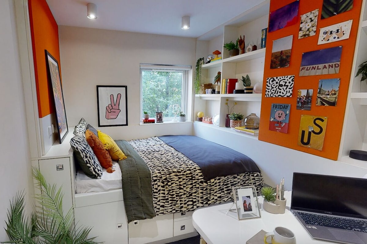 A modern bedroom with a bed, a desk with a laptop, a window, and a wall with various decorations.