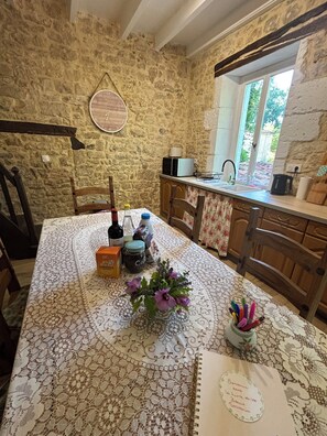 A rustic kitchen with a stone wall, a wooden table with a lace tablecloth, a clock, and a window with a view of greenery. - Cosy bright cottage perfect for families or some time in nature. Private pool! (Cellefrouin)