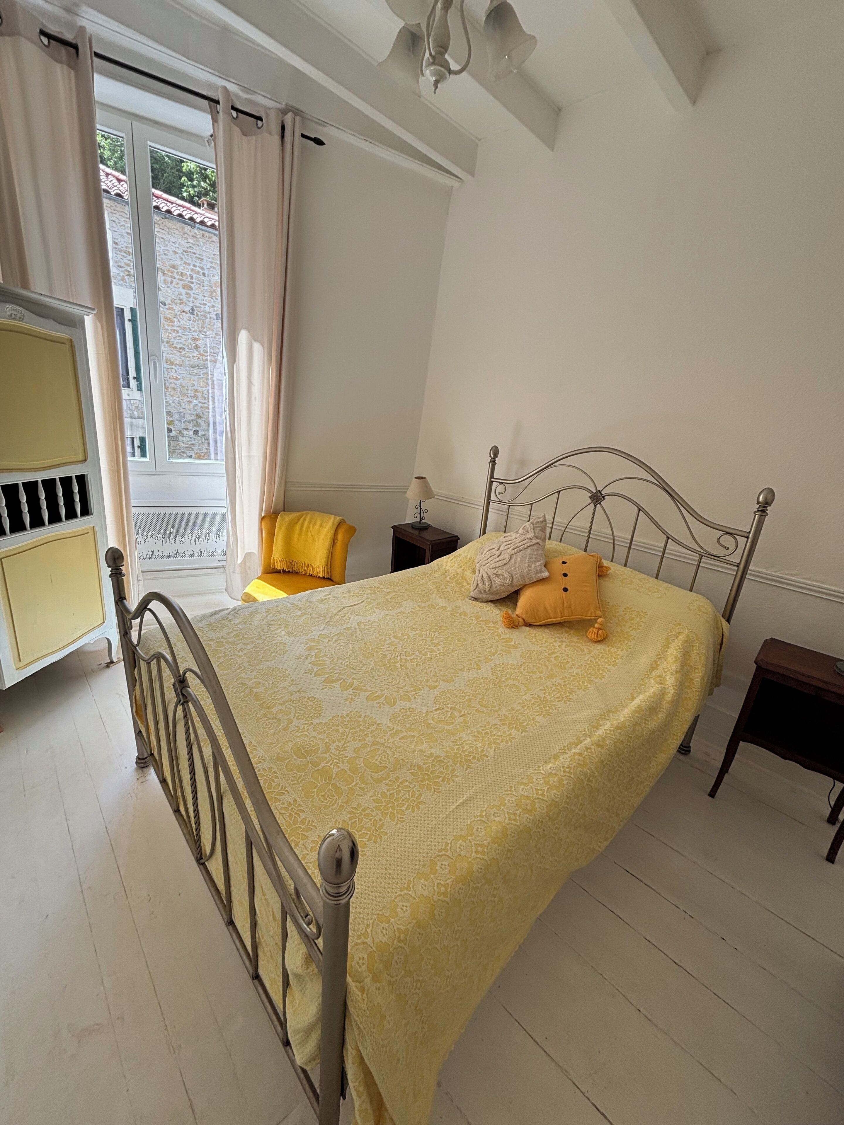 A bedroom with a metal bed frame, a yellow bedspread, a bedside table with a lamp, and a window with curtains.