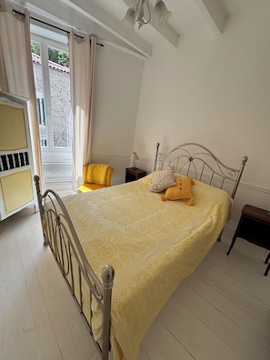A bedroom with a metal bed frame, a yellow bedspread, a bedside table with a lamp, and a window with curtains. - Cosy bright cottage perfect for families or some time in nature. Private pool! (Cellefrouin)
