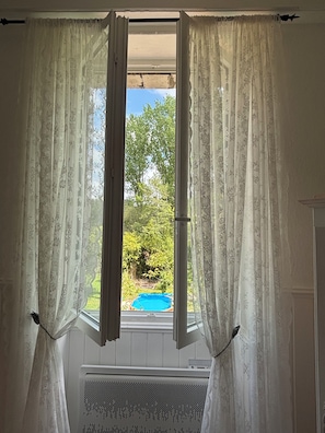 A window with white curtains, a view of greenery and a blue pool outside. - Cosy bright cottage perfect for families or some time in nature. Private pool! (Cellefrouin)
