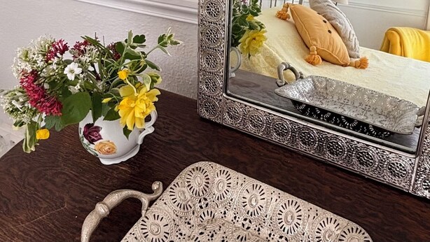Ein Spiegel mit aufwendigem Rahmen, ein Bett mit Metallkopfteil, ein Nachttisch mit Blumenvase und ein gemustertes Stuhl.