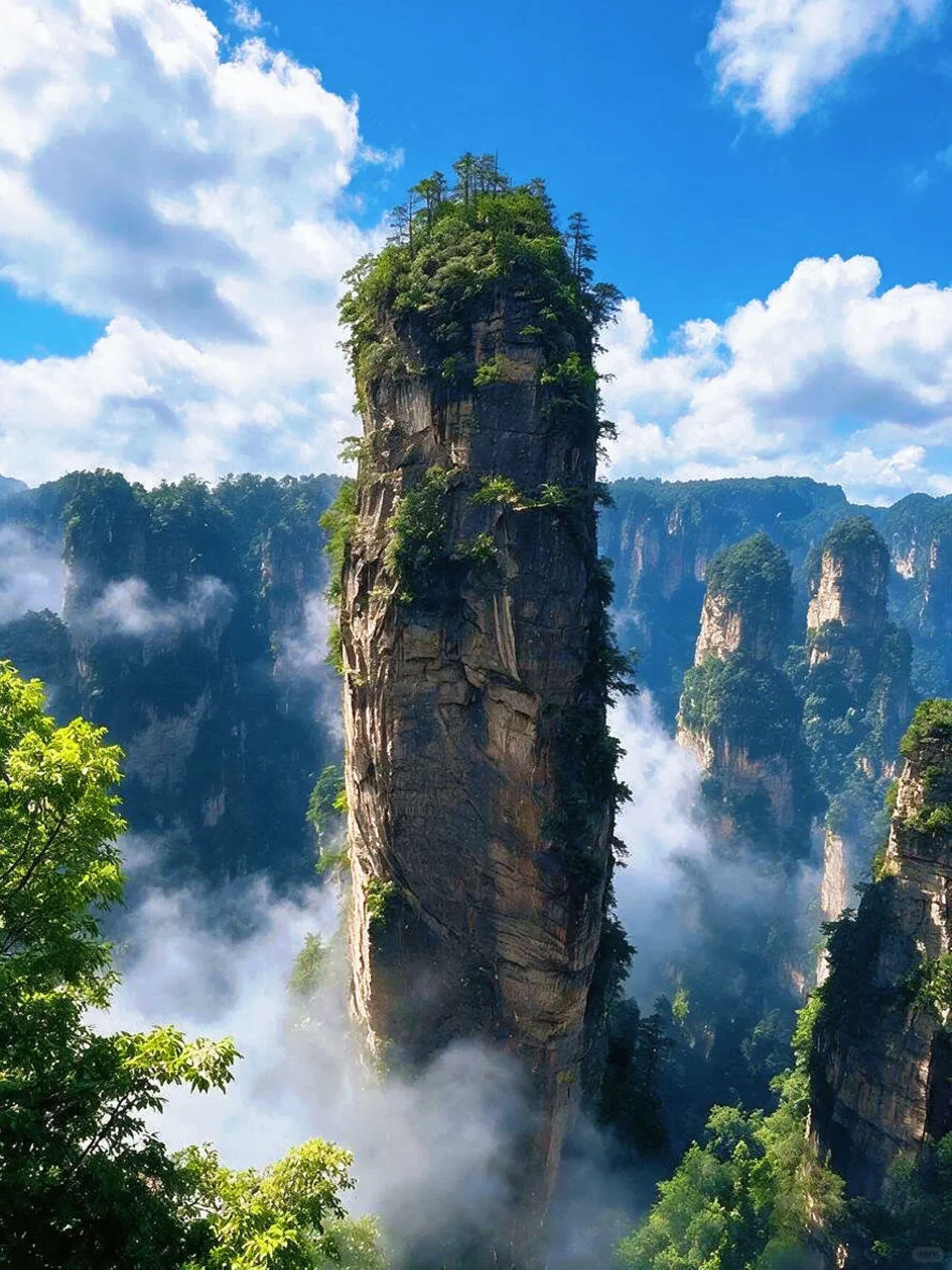 Formación rocosa alta y esbelta cubierta de vegetación densa, rodeada de montañas boscosas envueltas en niebla.