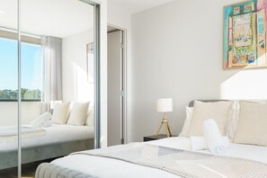 A hotel room with a bed, a bedside table with a lamp, a window with curtains, and a painting on the wall.