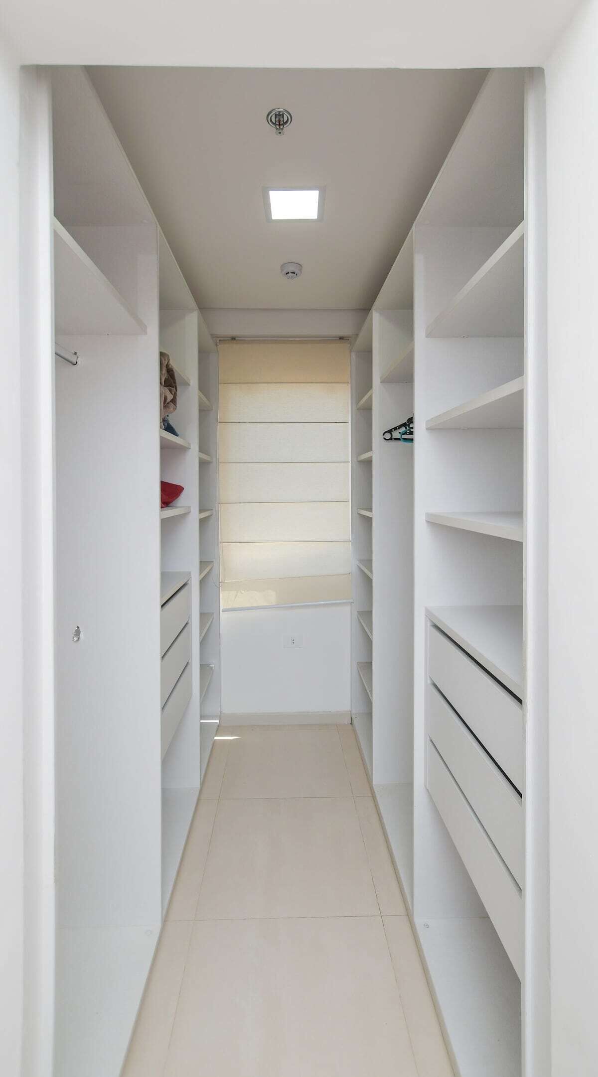 Un petit dressing étroit avec des étagères blanches, un luminaire au plafond et une grille d’aération.
