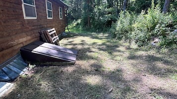 Une cabane rustique avec une façade en bois, une petite terrasse et une cour verdoyante entourée d’arbres.